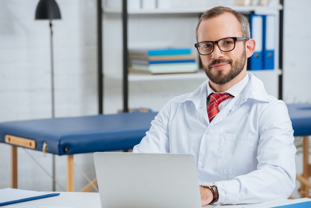 portrait of smiling male chiropractor in white coat at workplace with laptop in hospital